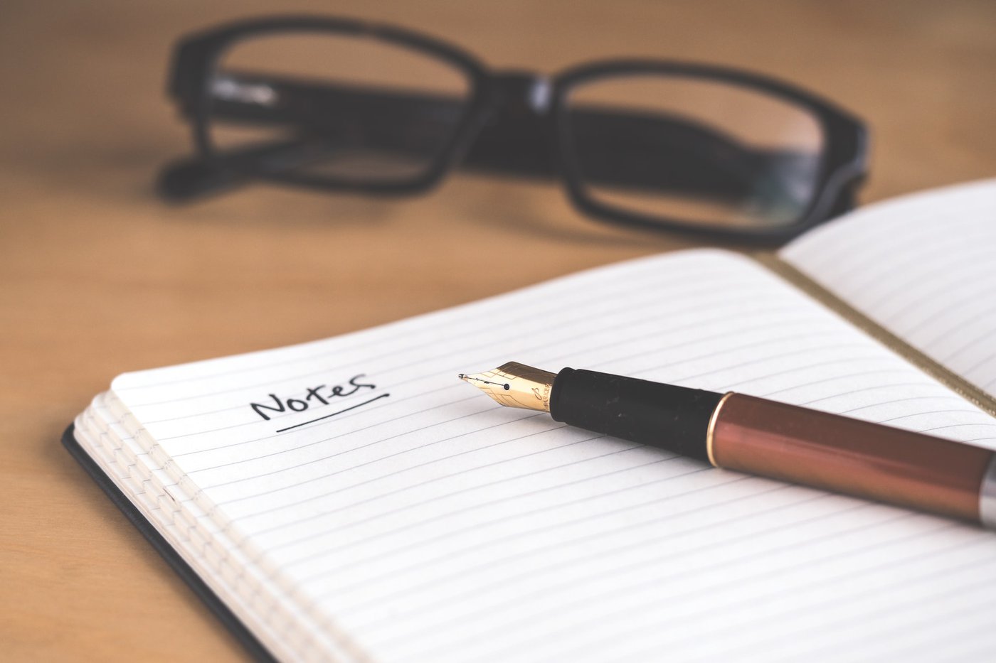 IMAGE DESCRIPTION: A notebook open with a pen sitting on top. The word "Notes" is written on the top of the page and underlined. Behind the notebook are a pair of reading glasses.