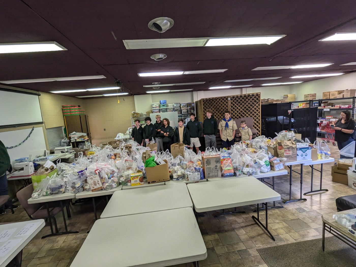 Nine boy scouts stand with bags of food spread across many tables.